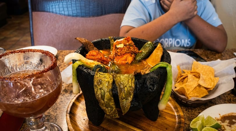 A molcajete filled with meat, peppers, and onions, alongside a drink and tortilla chips.