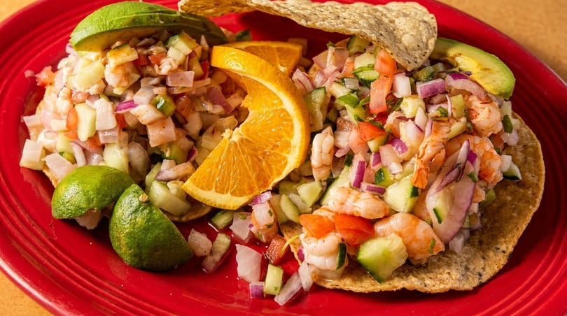 Two shrimp tostadas with diced vegetables, avocado, orange slices, and lime wedges on a red plate.