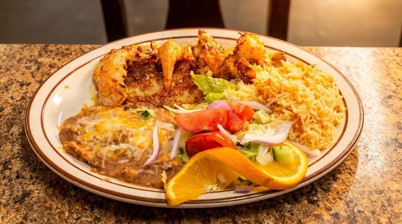 A plate of grilled shrimp, rice, refried beans with cheese, and a side salad with orange slices.